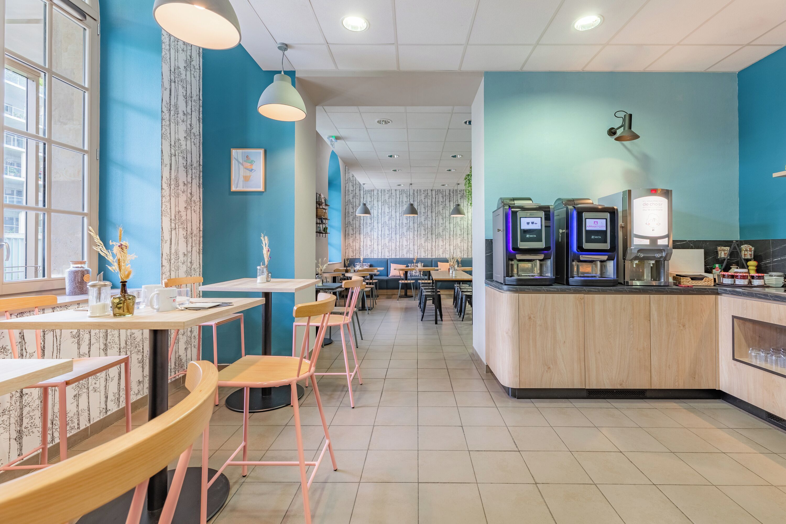 Een modern café met houten tafels en stoelen, een blauwe accentmuur en zelfbedieningskoffiemachines.