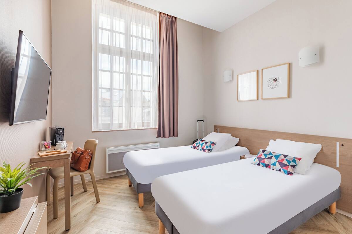A hotel room with two beds, a desk with a coffee maker, a TV, and a window with curtains.