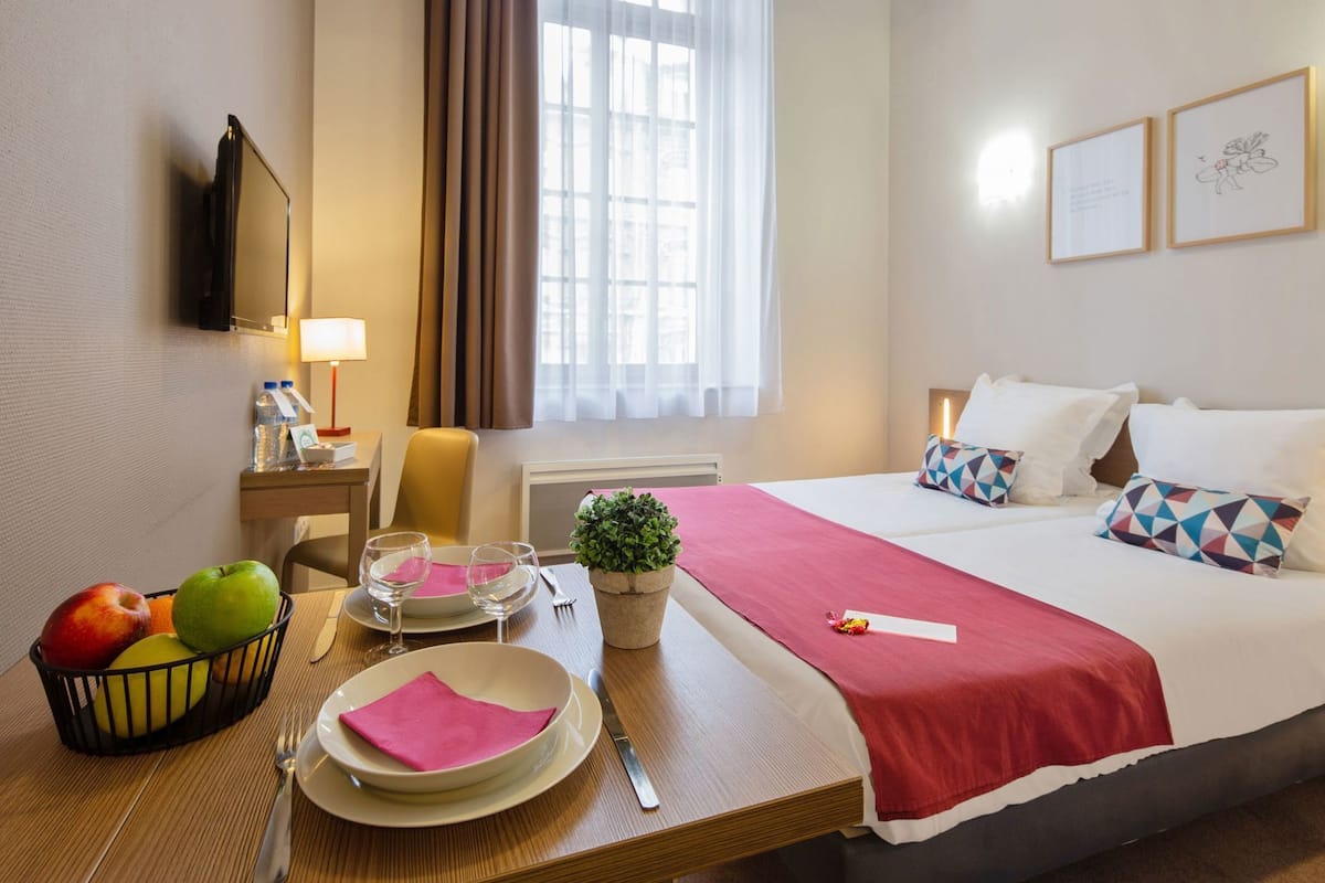 A hotel room with a bed, bedside table, lamp, TV, window with curtains, and a small plant.