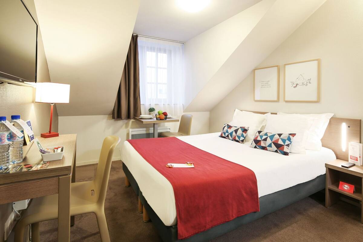 A hotel room with a bed, desk, chair, and a small table with a lamp.