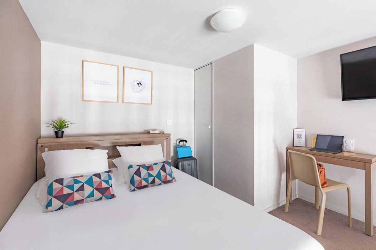 A hotel room with a bed, a desk with a laptop, a chair, a suitcase, and a TV on the wall.