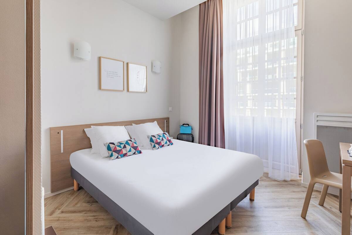 A neatly made bed with pillows, a bedside table, a chair, and a window with curtains.