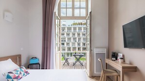 A hotel room with a bed, a desk, a chair, a suitcase, a table with chairs, and a balcony with a view of buildings. - Appart'City Confort Reims Centre (Reims)