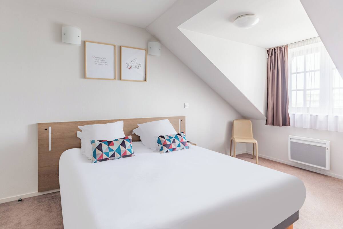 A modern hotel room with a large bed, a wooden headboard, two framed artworks, a bedside table, a chair, and a window with curtains.