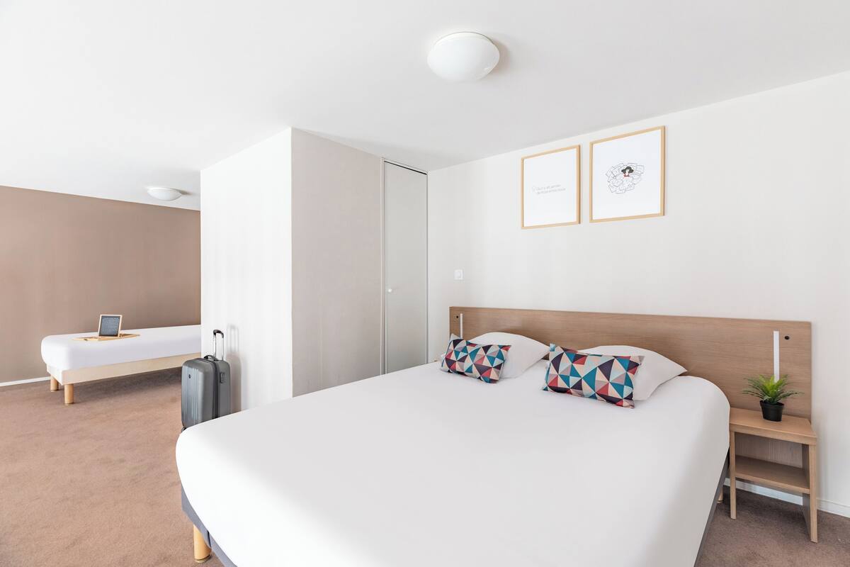 A modern hotel room with a large bed, a bedside table with a plant, and a desk with a laptop.