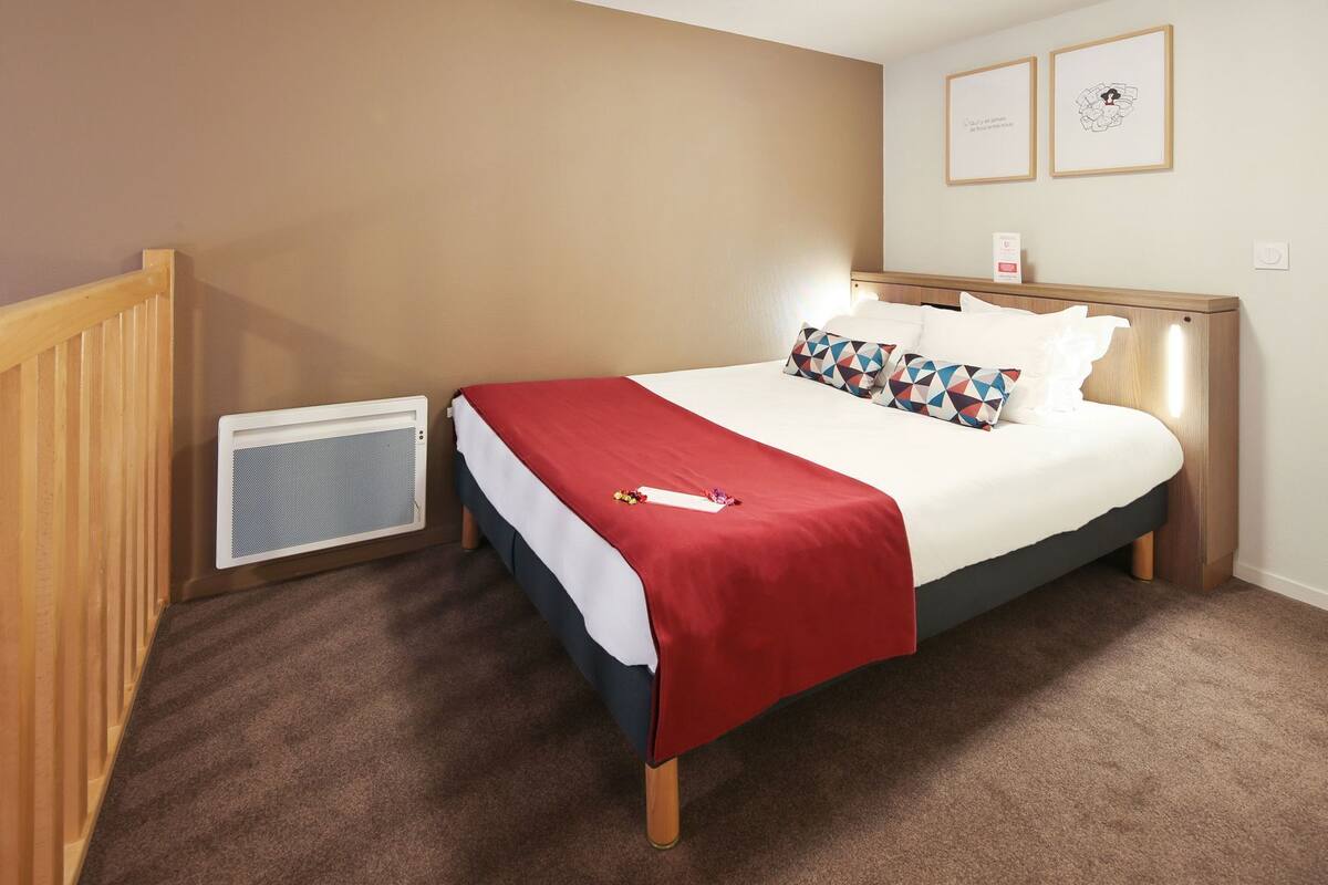 A neatly arranged hotel room with a bed, a heater, and two framed pictures on the wall.