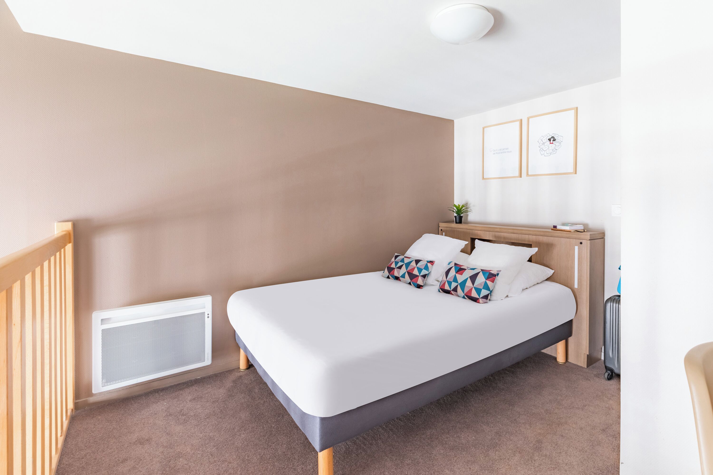 Ein ordentlich eingerichtetes Hotelzimmer mit einem Bett, einem kleinen Nachttisch, einem Wandlüfter und zwei gerahmten Bildern an der Wand.