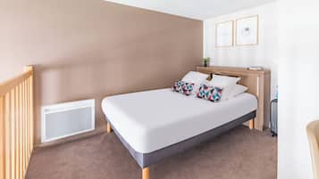 Ein ordentlich eingerichtetes Hotelzimmer mit einem Bett, einem kleinen Nachttisch, einem Wandlüfter und zwei gerahmten Bildern an der Wand.