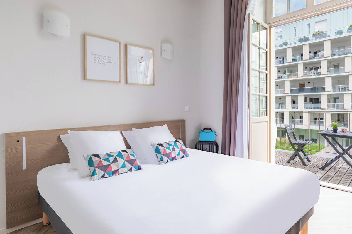 A hotel room with a bed, two framed pictures on the wall, a window with a view of the building, and a balcony with a table and chairs.