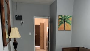 Un salón con un sofá marrón, una mesa de centro de vidrio y una lámpara. Hay una puerta que conduce a otra habitación y un cuadro de una palmera en la pared.