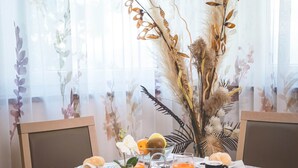 A breakfast table set with various dishes, including pastries, fruit, and beverages, in a room with sheer curtains and a decorative plant arrangement. - Hotel Fai (Fai della Paganella)