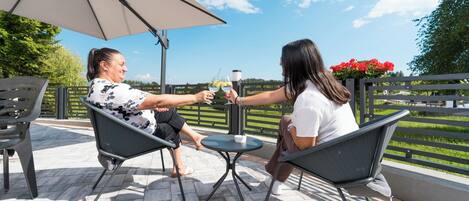 Dos personas sentadas en una mesa bajo un gran paraguas, en un patio con vista a un paisaje verde.