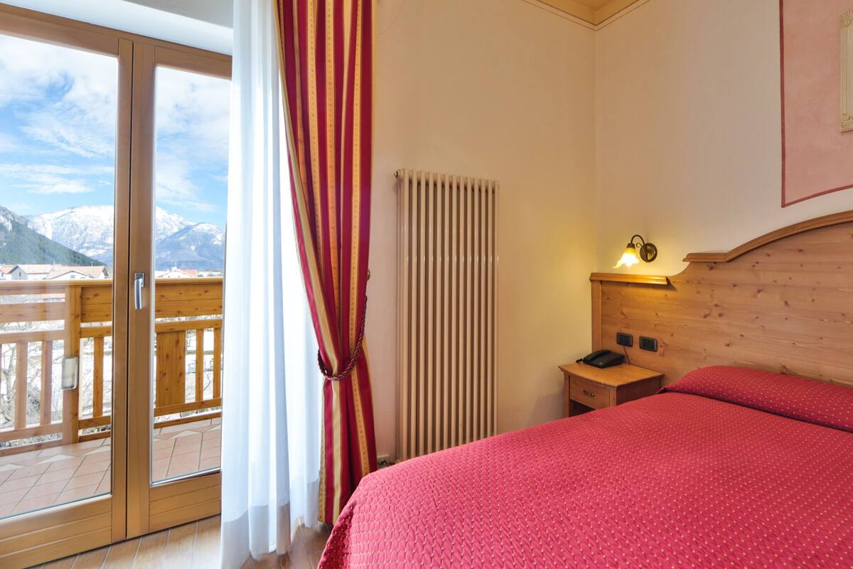 A bedroom with a bed, a nightstand, a lamp, a radiator, and a view of mountains through a sliding glass door.