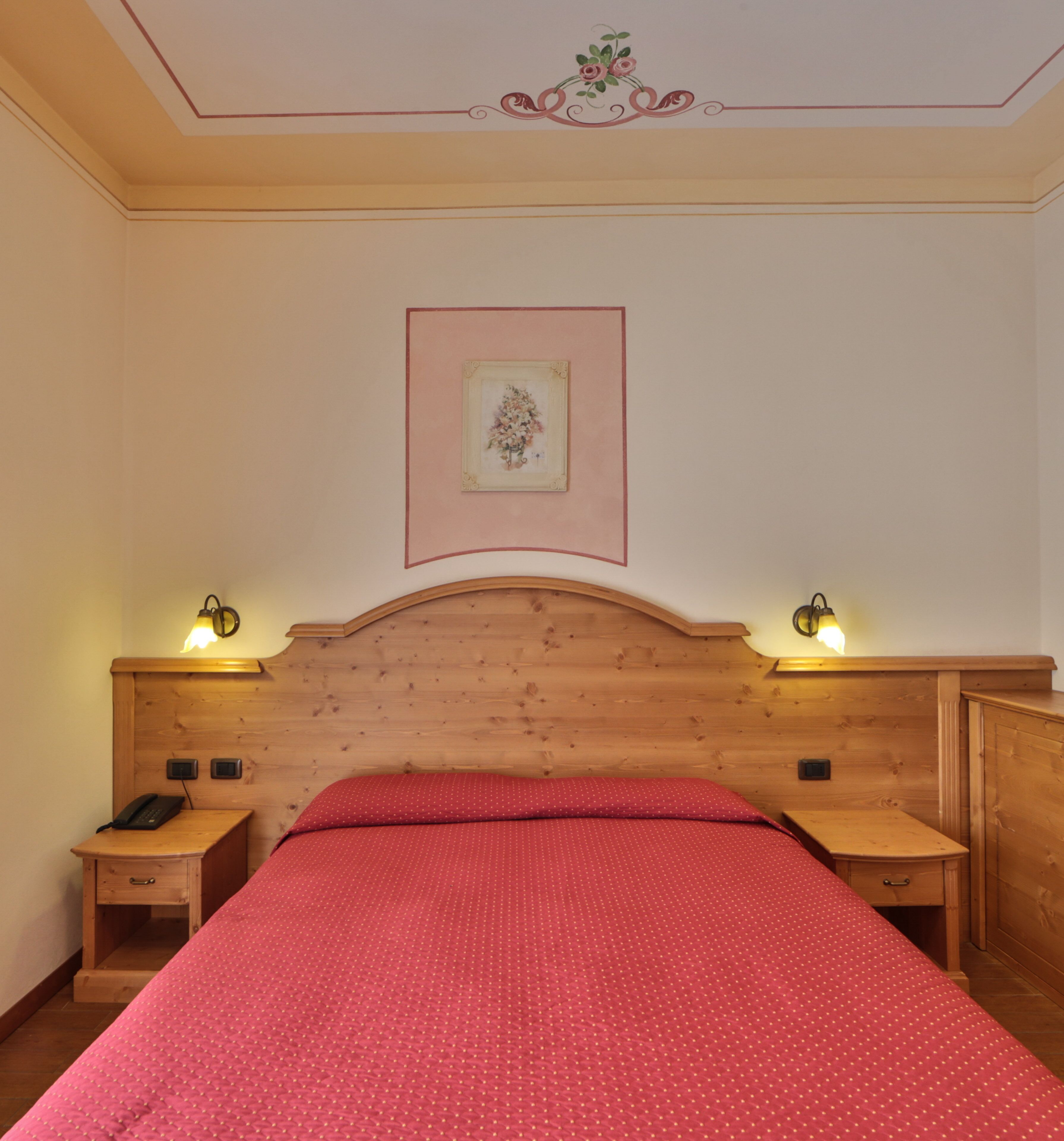Quarto com cabeceira de madeira, colcha vermelha, duas mesas de cabeceira com abajures e um quadro na parede.