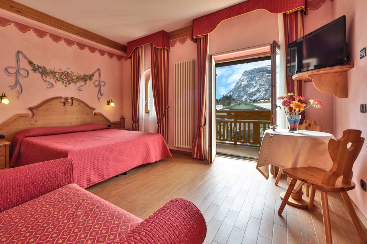 A hotel room with two beds, a balcony with a mountain view, a TV, and a table with a vase of flowers.