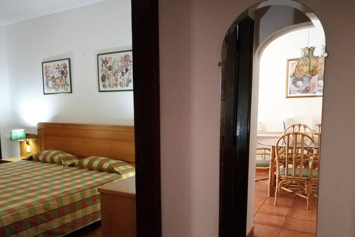 A bedroom with a bed, a nightstand, and a wall-mounted lamp. There is a dining area with a table and chairs visible through an archway.