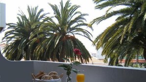 A balcony with a table set for breakfast, including bread, fruit, and a cup of coffee, overlooking palm trees and a building. - Solar de São João - Apartamentos Turísticos (Albufeira)