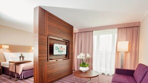 Habitación de hotel con cama, escritorio con computadora, sofá, televisor montado en un panel de madera y ventana con cortinas.