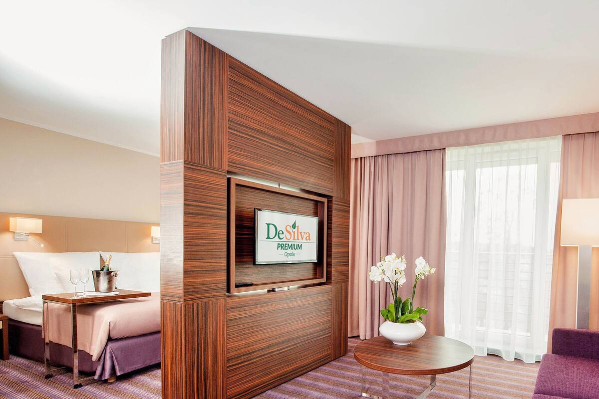 Ein Hotelzimmer mit einem Bett, einem Schreibtisch mit Computer, einer Couch, einem an einer Holzwand montierten Fernseher und einem Fenster mit Vorhängen.