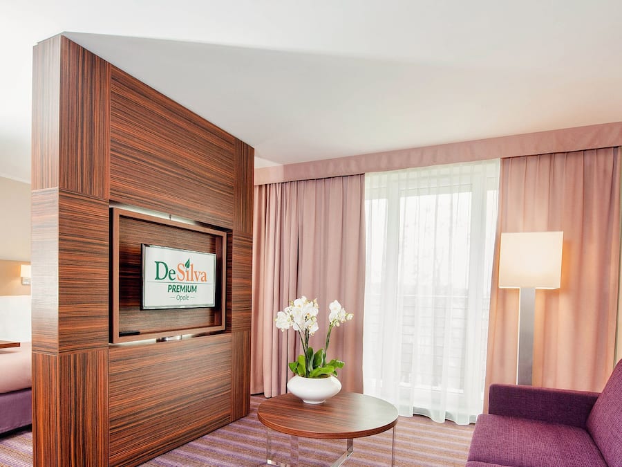 Een hotelkamer met een bed, een bureau met een computer, een bank, een televisie bevestigd aan een houten paneel, en een raam met gordijnen.