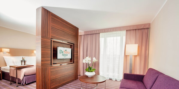 A hotel room with a bed, a desk with a computer, a sofa, a TV mounted on a wooden panel, and a window with curtains. - DeSilva Premium Opole (Opole)