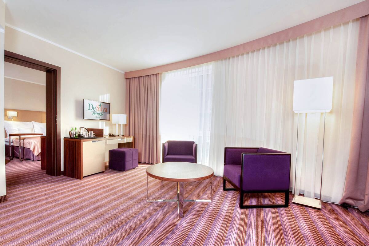 Ein Hotelzimmer mit einem Bett, einem Schreibtisch mit Computer, einem violetten Sessel, einem runden Tisch und einem großen Fenster mit Vorhängen.