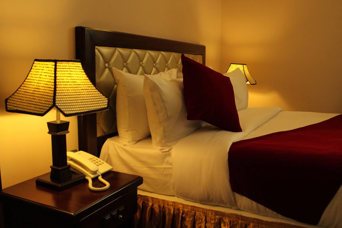 A hotel room with a bed, a nightstand with a lamp, a telephone, and a red throw blanket.