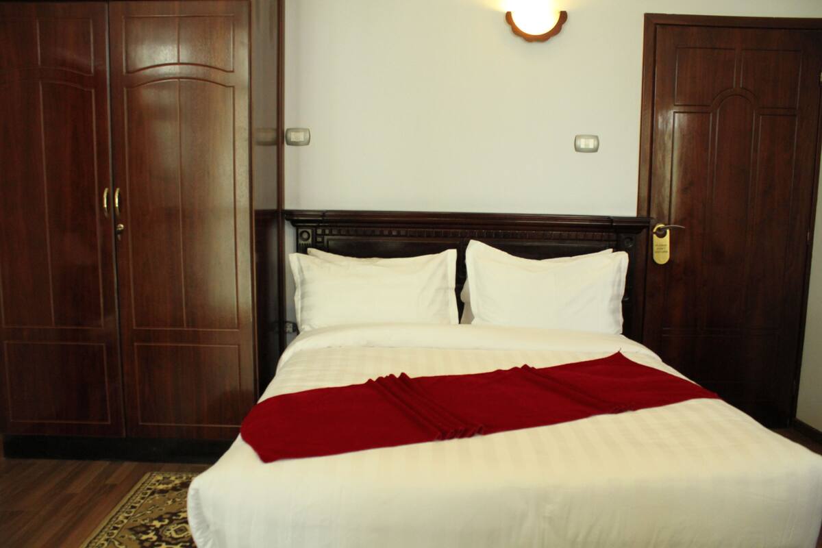 A hotel room with a large bed, wooden wardrobe, and a wall-mounted light fixture.