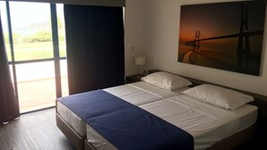 A hotel room with a large bed, a nightstand, a lamp, and a painting of a sunset over a bridge. - Almar Hotel Apartamento (Albufeira)