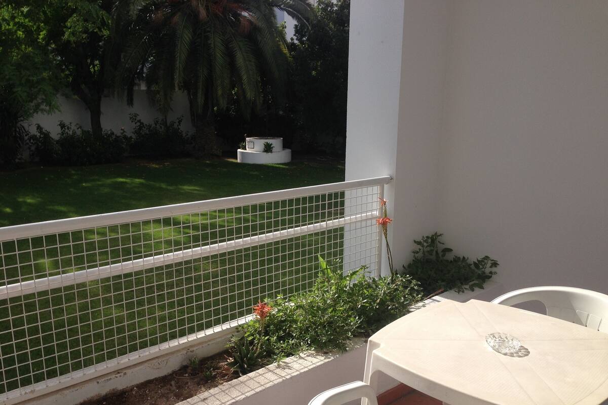 Un balcón con mesa y sillas blancas, barandilla blanca, y vistas a un césped verde y palmeras.