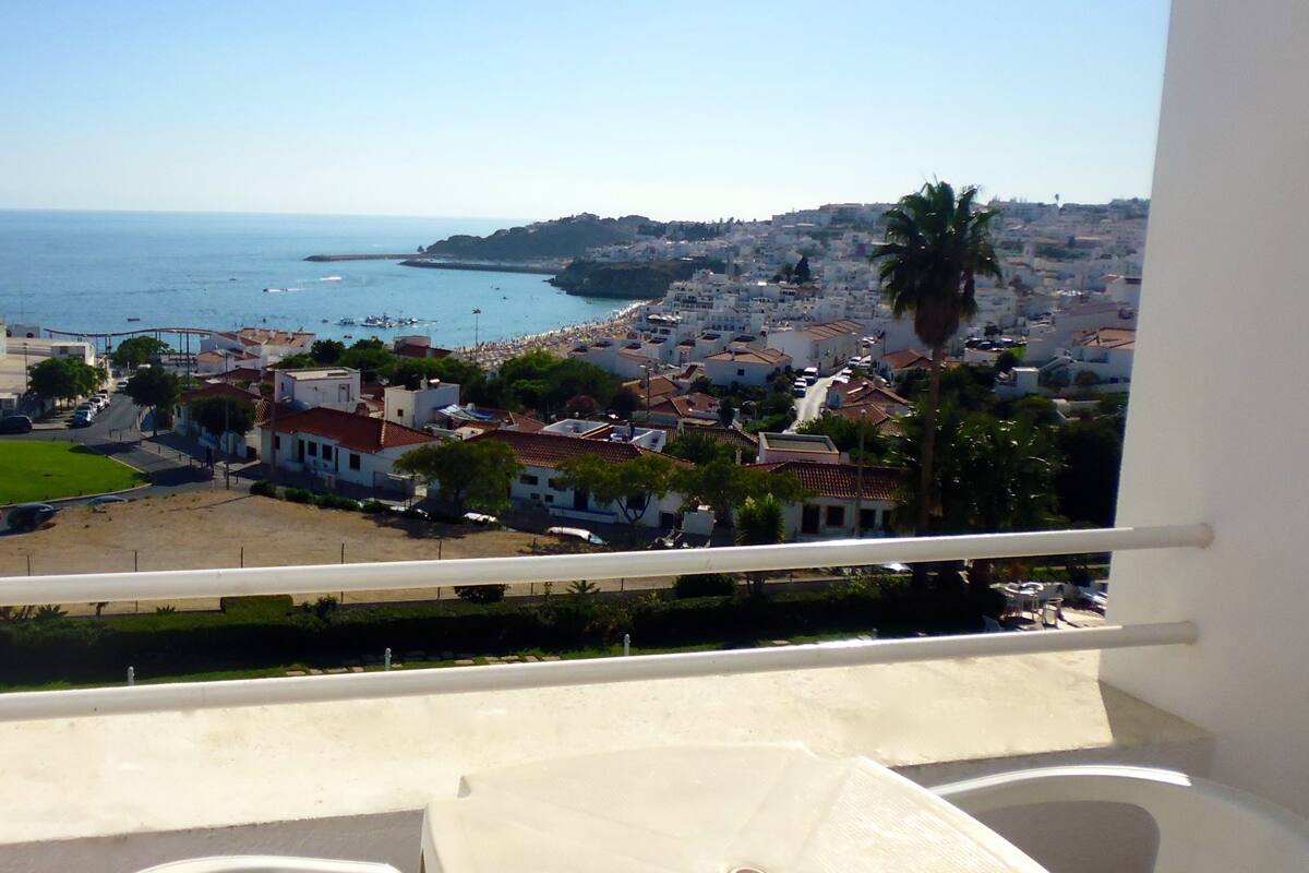 Un balcón con mesa y sillas, con vista a un pueblo costero, edificios, una playa y el mar.