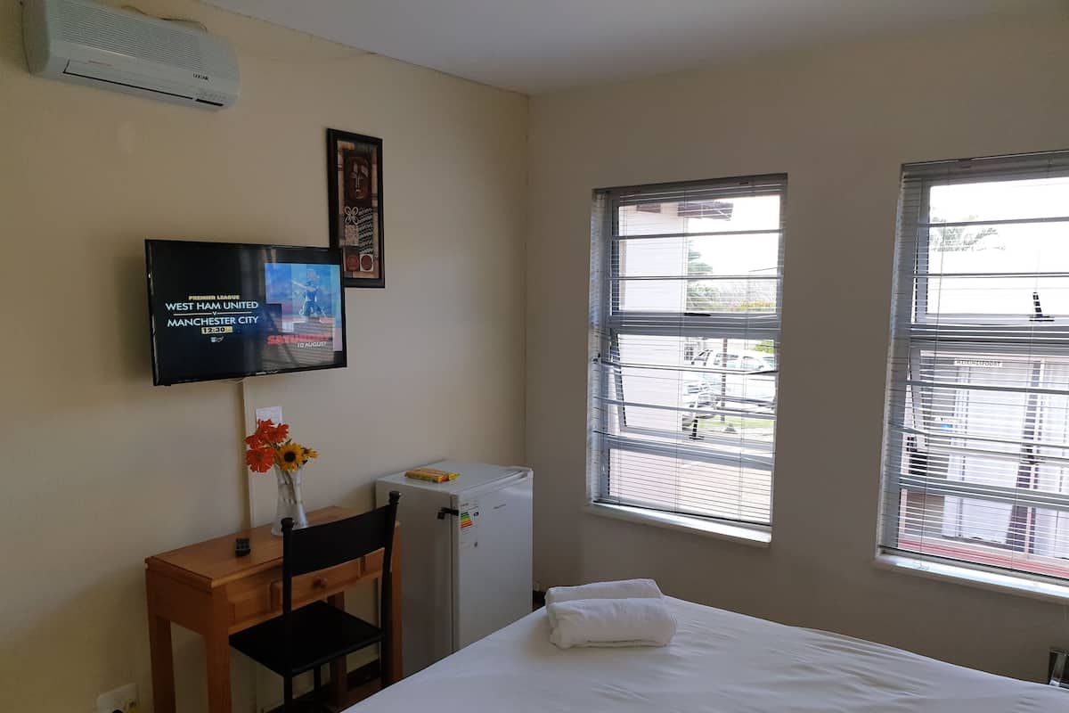 A hotel room with a bed, a desk with a chair, a television mounted on the wall, and a window with blinds.