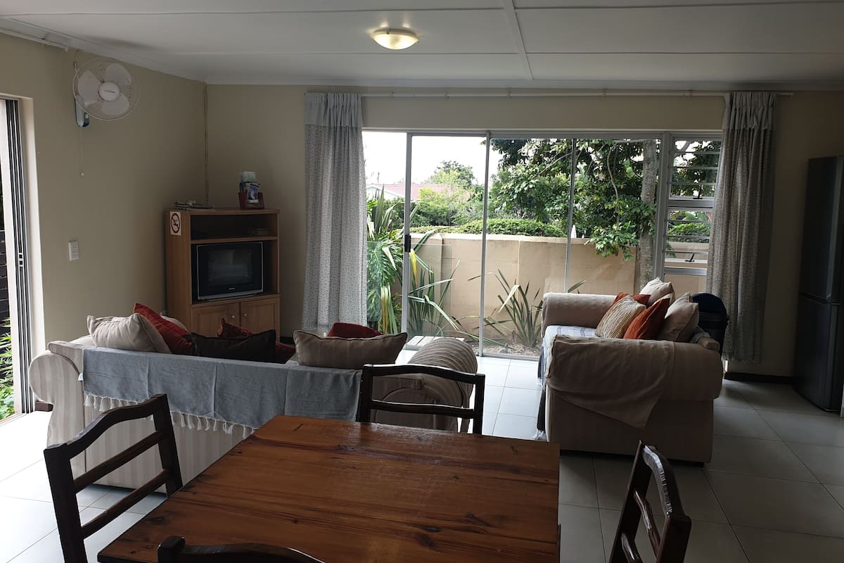 A living room with a wooden dining table, two sofas, a TV on a wooden cabinet, and a sliding glass door leading to a balcony with plants.