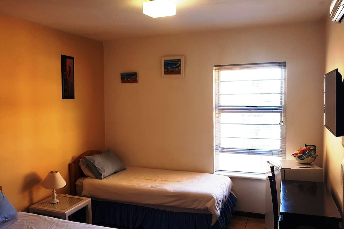 A small, single-bedroom room with two beds, a desk, a lamp, and a window with blinds.