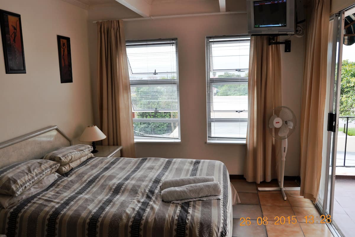A hotel room with a bed, a TV, a fan, and two windows with curtains.