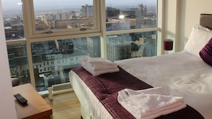 A hotel room with a large window offering a city view, a neatly made bed, and a remote control on a small table. - Glasgow Lofts (Glasgow)
