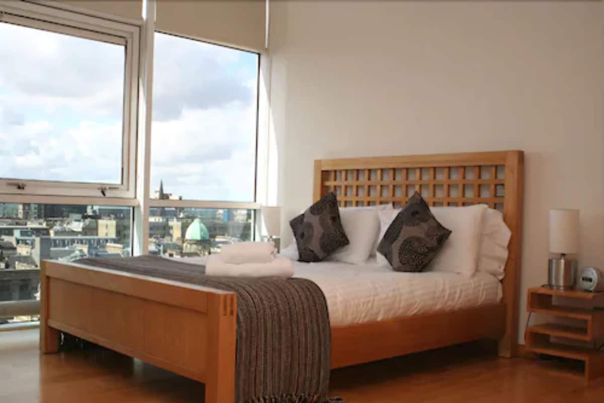 A bedroom with a wooden bed, a view of the city through large windows, and a nightstand with a lamp.
