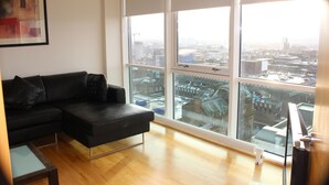 A modern living room with a black leather sofa, a glass coffee table, and a view of a cityscape through large windows. - Glasgow Lofts (Glasgow)
