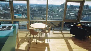 A modern room with a large window, a round table with chairs, a sofa, and a kitchen area with a stove. - Glasgow Lofts (Glasgow)