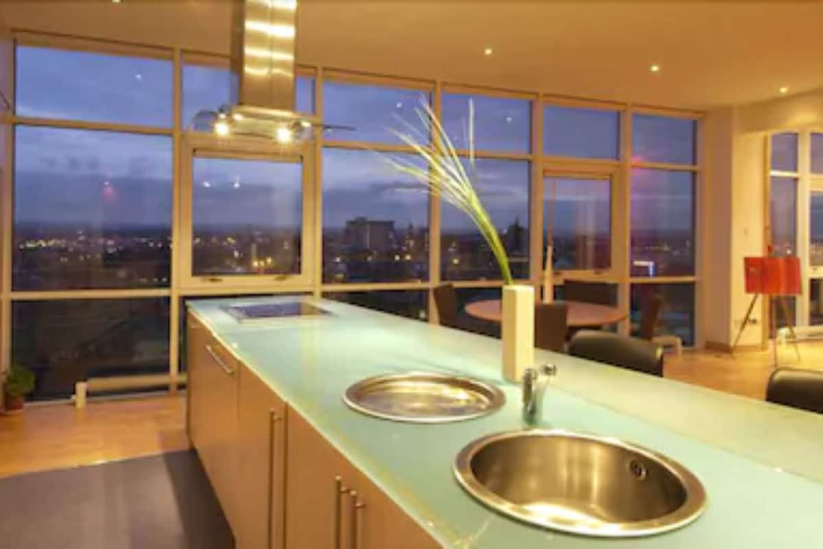 A modern kitchen with a large window, a stainless steel sink, and a view of the city at dusk.