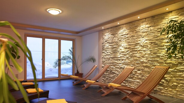 A spa area with wooden loungers, a stone wall, and a large window with a view. - Haus Anna Apartments (Klösterle am Arlberg)