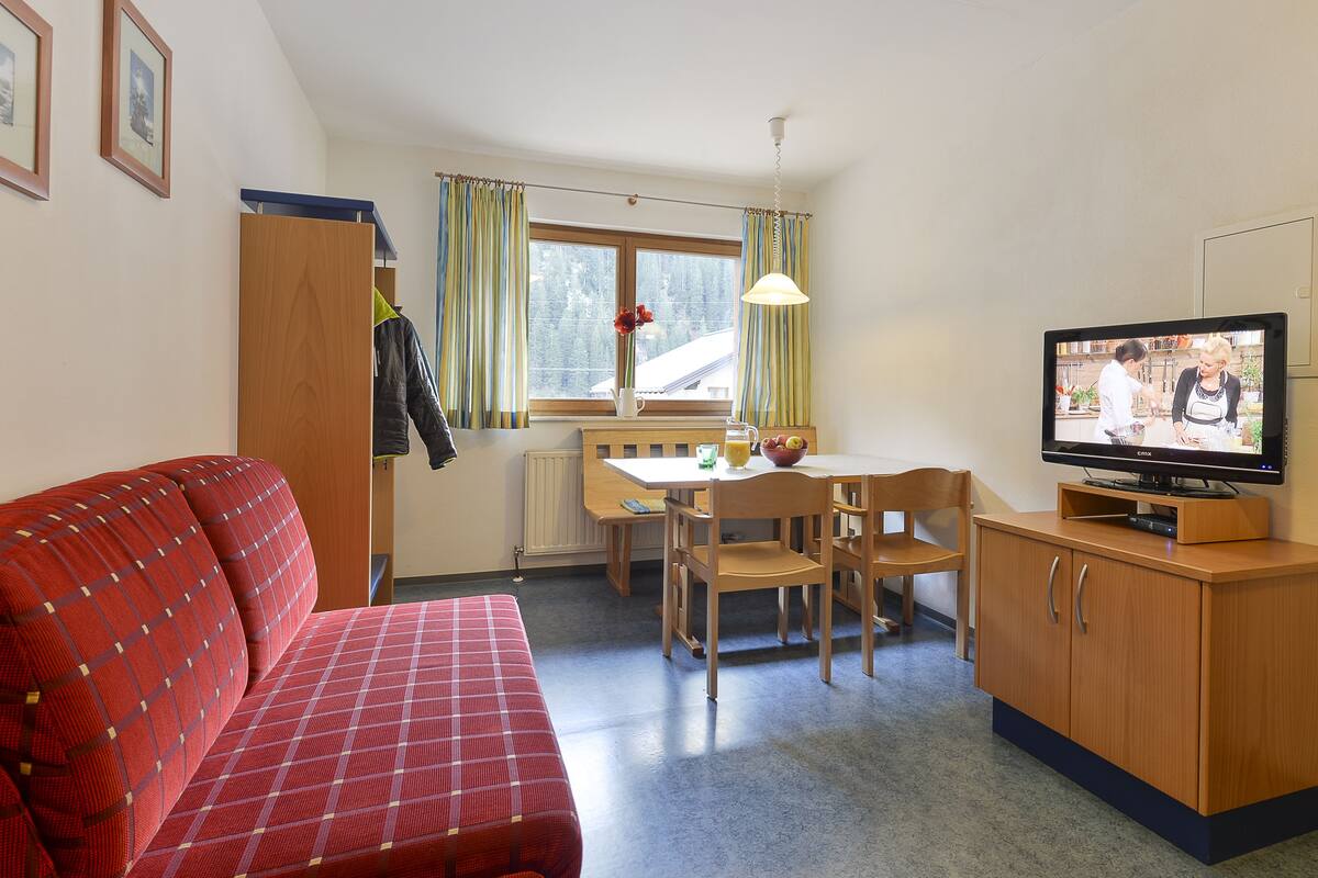 A compact room with a red sofa, wooden dining table, chairs, a TV, and a small kitchen area.