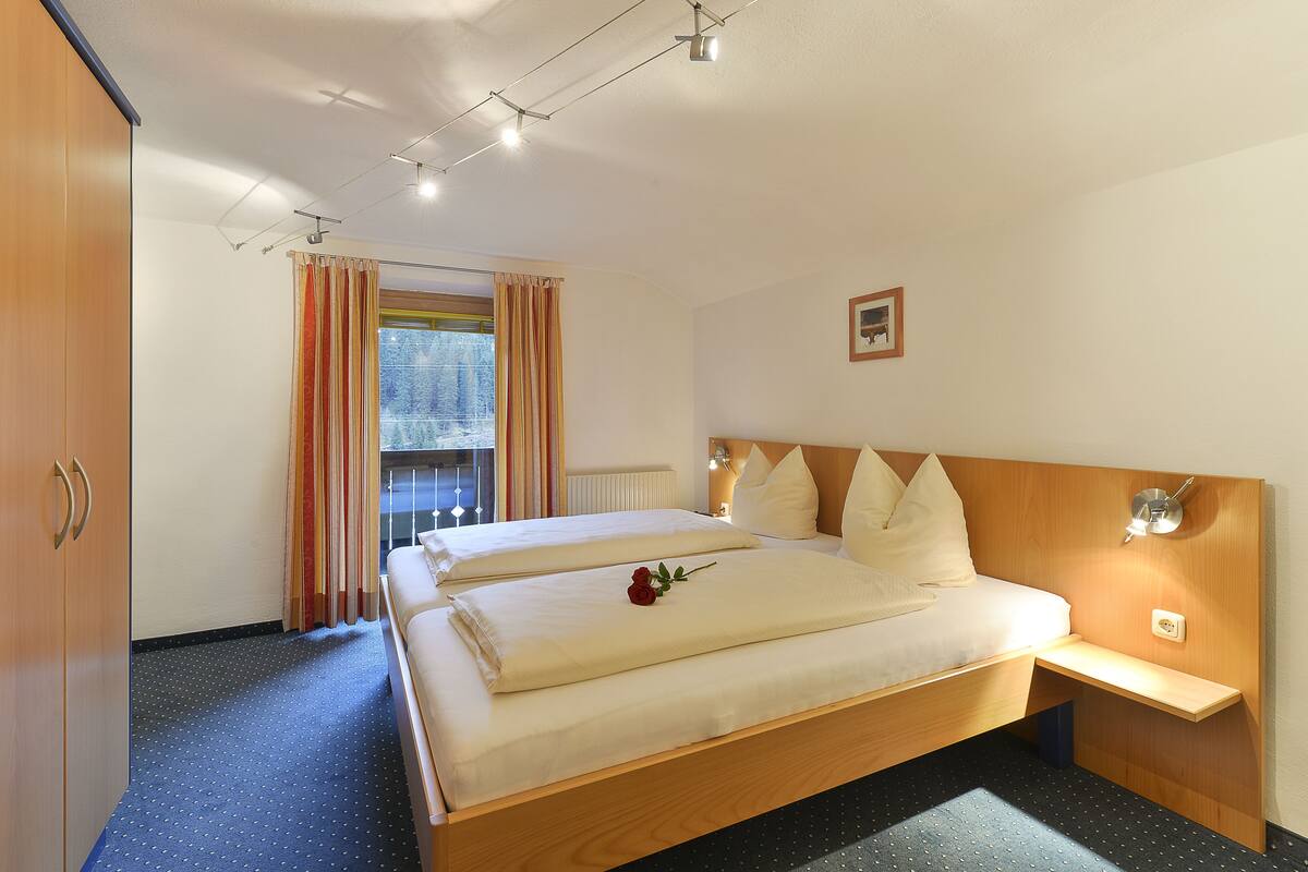 A hotel room with a large bed, a bedside table, a nightstand, a window with curtains, and a view of trees outside.