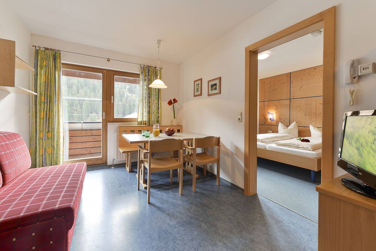 A compact hotel room with a bed, a dining table with chairs, a TV, and a bathroom visible through an open door.