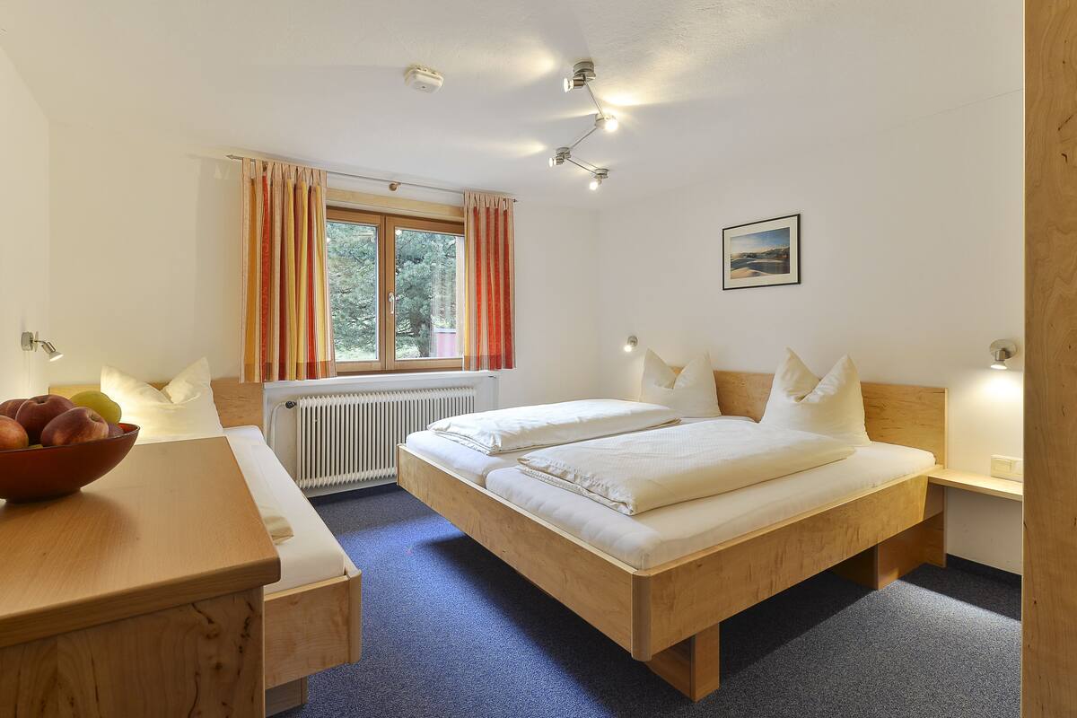 A bedroom with a wooden bed, a bedside table with a bowl of fruit, a window with curtains, and a radiator.