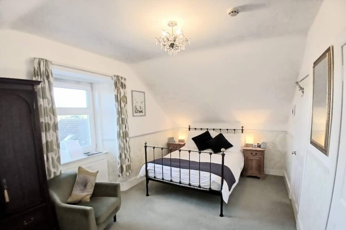A bedroom with a bed, a chair, a nightstand, a dresser, a window with curtains, a chandelier, and a mirror.