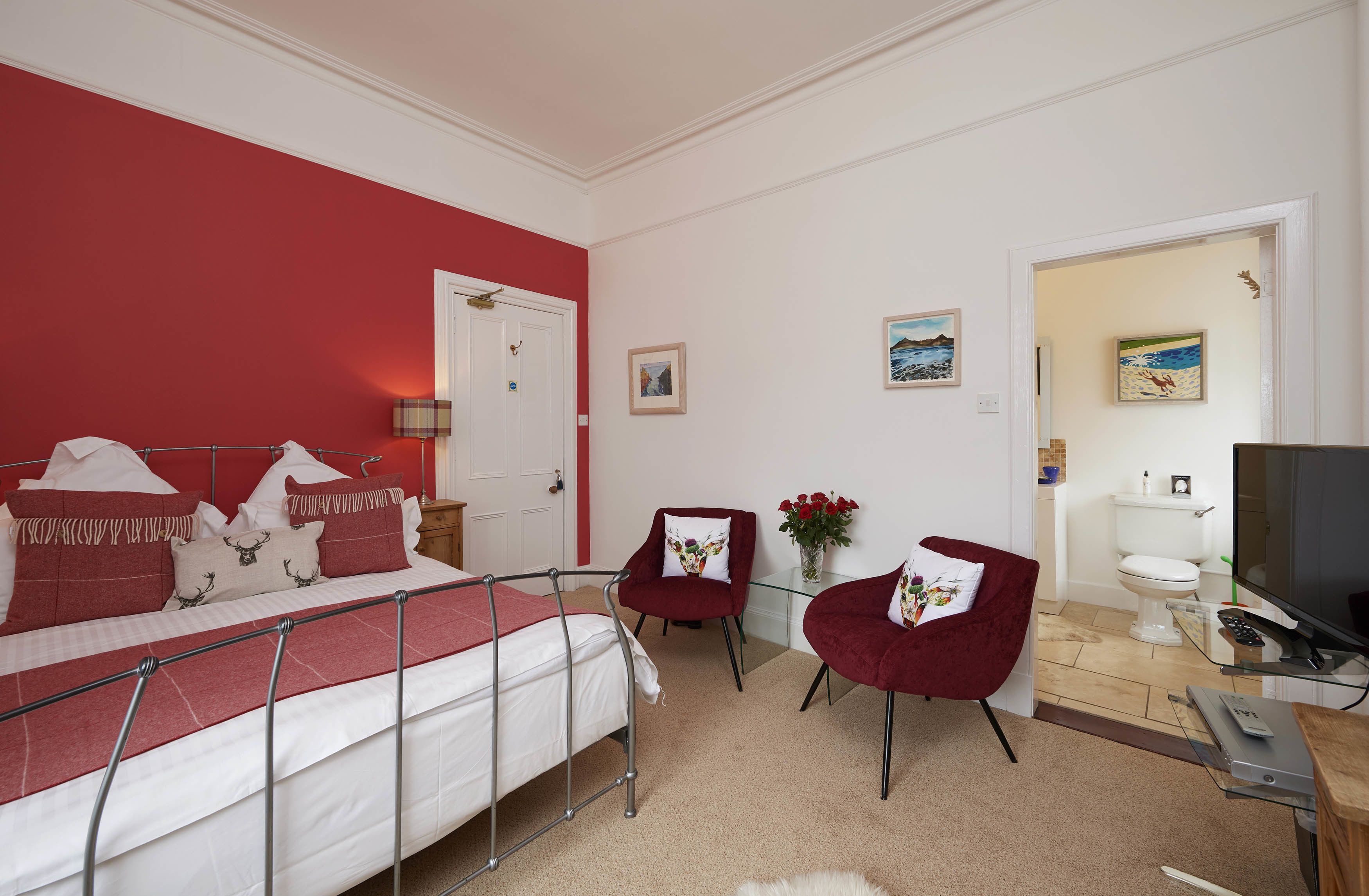 Ein Schlafzimmer mit einem Bett, zwei roten Stühlen, einem Fernseher und einem durchgegangenen Tür erkennbaren Badezimmer.