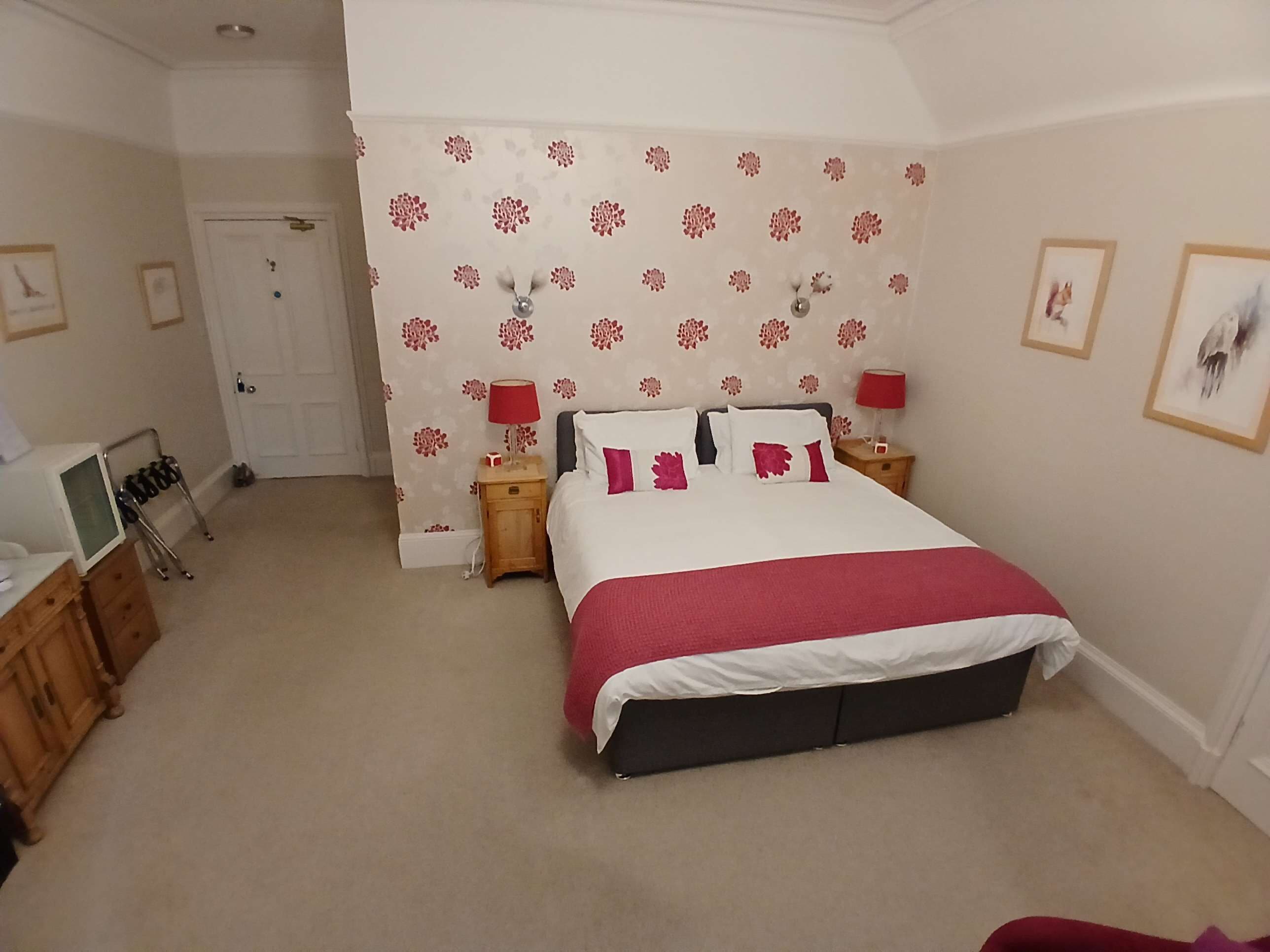 A bedroom with a bed, two bedside tables, a wooden dresser, a door, and a wall with floral wallpaper.