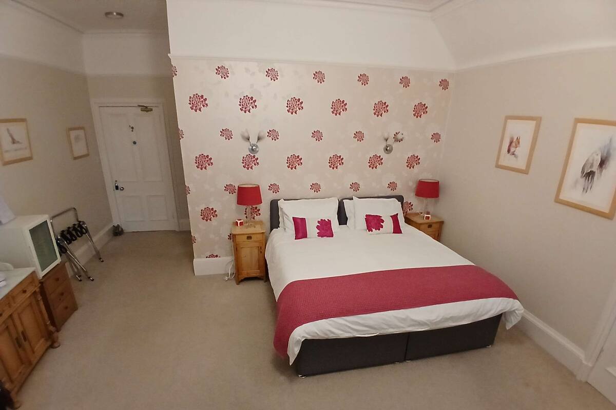 A bedroom with a bed, two bedside tables, a wooden dresser, a door, and a wall with floral wallpaper.