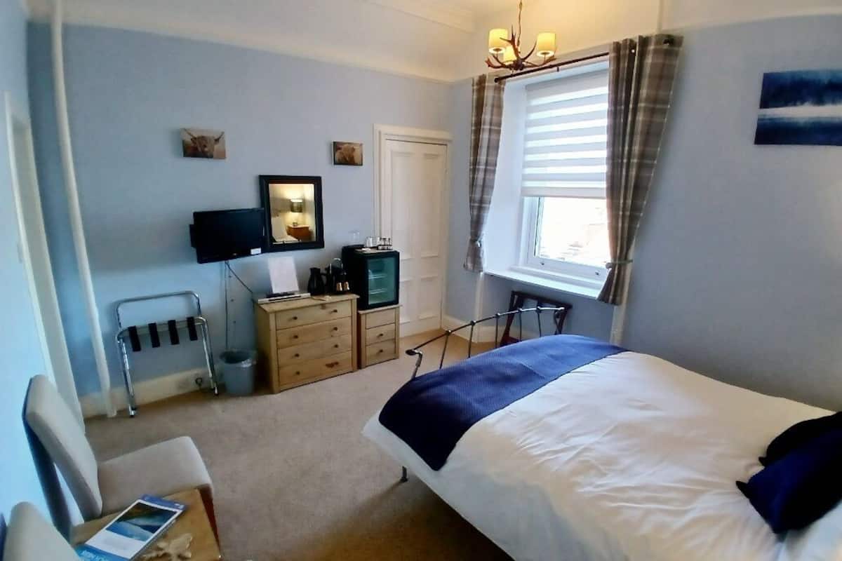 A bedroom with a bed, a desk, a chair, a television, and a window with curtains.